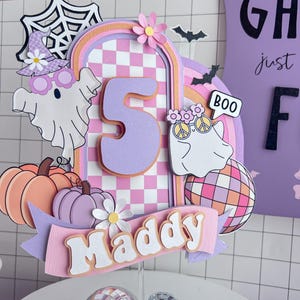 May include: A layered paper cake topper featuring a large number 5, the name Maddy, two ghosts, pumpkins, a disco ball, and a spiderweb. The design uses a pastel color palette of purple, pink, and orange with a checkered pattern and floral accents.
