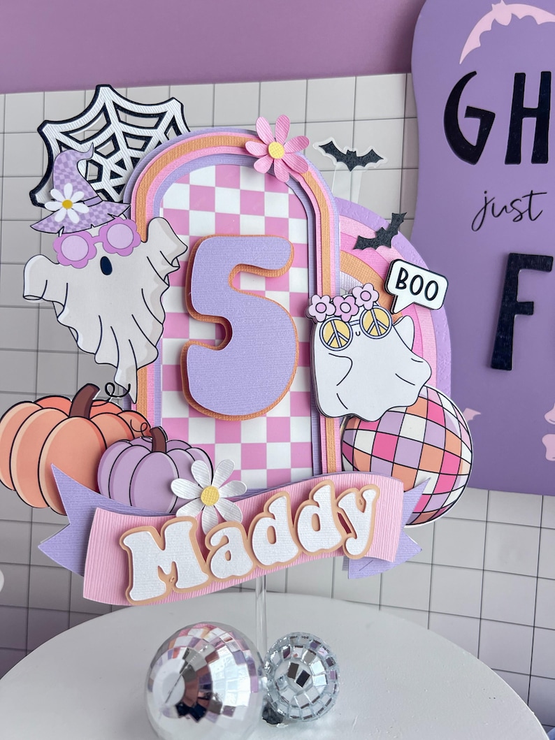 May include: A layered paper cake topper featuring a large number 5, the name Maddy, two ghosts, pumpkins, a disco ball, and a spiderweb. The design uses a pastel color palette of purple, pink, and orange with a checkered pattern and floral accents.