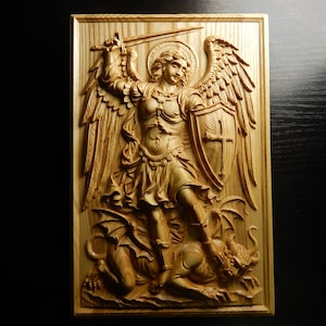 May include: A wooden relief carving of Saint Michael the Archangel, shown with wings, a sword, and a shield bearing a cross. The angel is depicted standing over a defeated demon. The carving is framed in a rectangular shape.