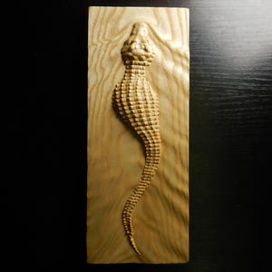 Könnte beinhalten: Holzschnitzerei eines Seepferdchens. Das Seepferdchen ist als Relief auf einer rechteckigen Holztafel geschnitzt. Das Seepferdchen ist hellbraun und hat eine strukturierte Oberfläche. Die Holzmaserung ist im Hintergrund sichtbar.