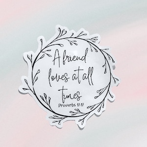 May include: White sticker with black script that reads "A friend loves at all times" surrounded by a black floral wreath. The text "Proverbs 17:17" is below the quote. The background is a gradient of pink and light blue.