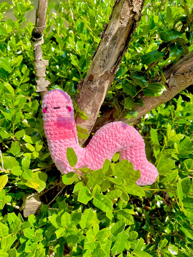 May include: A pink crocheted worm plush toy with a smiling face. The worm is curled up in a bush with green leaves.