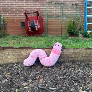 May include: A pink and white plush worm toy with a smiling face. The worm is lying on a bed of brown mulch.