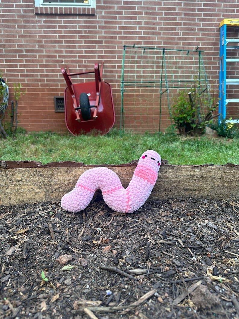 May include: A pink and white plush worm toy with a smiling face. The worm is lying on a bed of brown mulch.