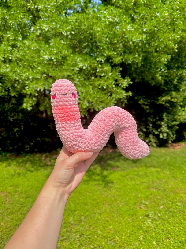 May include: A pink crocheted worm plush toy with a smiling face and pink cheeks. The worm is being held in a hand against a green grassy background.