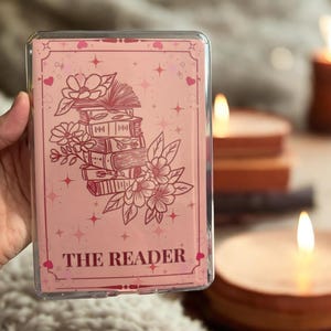 May include: A pink phone case with a floral and book design. The case features a stack of books, flowers, and the words "THE READER" in a decorative font. The case is held in a hand.