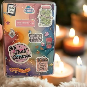 May include: A clear phone case adorned with various stickers. The stickers display text such as "Kindle Girlie," "Go Away I'm Reading," and "No Shelf Control Book Club." Other stickers show a jar of money, a coffee cup, and a tote bag filled with books. The background has a watercolor effect.