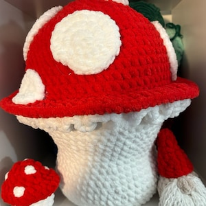 May include: A crocheted mushroom hat with a white base and a red top with white spots. There is a smaller crocheted mushroom with white spots next to it.