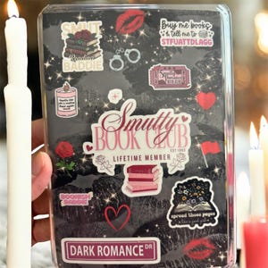 May include: A black book cover with various stickers, including a stack of books, a candle, and the words "Smutty Book Club." Other stickers include a red heart, a pair of handcuffs, and the text "Dark Romance DR."