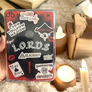 May include: A clear phone case with a black and red design, featuring stickers with text including "LORDS", "BLACKOUT", and "CARNAGE". The case is held in a hand, with candles and books in the background.