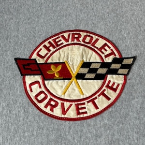 May include: A circular embroidered patch featuring the Chevrolet Corvette logo. The design includes red text reading CHEVROLET CORVETTE, a red flag with a gold fleur-de-lis, and a black and white checkered flag crossed over a white background.