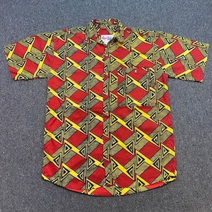 May include: A short-sleeve button-down shirt featuring a repeating geometric pattern of red, olive green, and yellow triangles with black outlines. The shirt includes a single chest pocket on the left side and a white label with the text ibis.