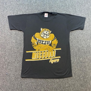 Puede incluir: Una camiseta negra de manga corta con un gráfico de un tigre que lleva una camiseta amarilla con el texto MIZZOU. Debajo del tigre, el texto MISSOURI está impreso en letras amarillas grandes entre dos líneas horizontales, seguido de Tigers.