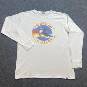 May include: A white long-sleeve t-shirt featuring a circular graphic on the chest. The graphic depicts a skier in motion with the text LOVELAND SKI AREA COLORADO in red letters. The design includes horizontal stripes of red, orange, and blue.