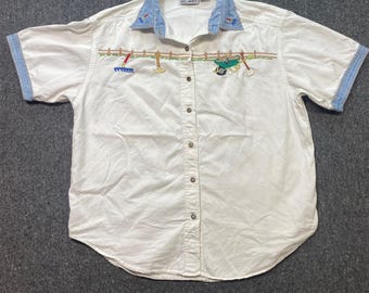 Könnte beinhalten: Ein weißes, kurzärmeliges Hemd mit Knopfleiste, hellblauem Jeanskragen und Ärmelbündchen. Das Hemd zeigt gestickte Gartengeräte wie eine Harke, eine Hacke und eine Schubkarre entlang einer Zaunlinie im Brustbereich.