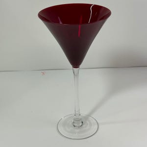 May include: A martini glass featuring a deep red, conical bowl and a slender, clear glass stem that connects to a circular, transparent base. The design is sleek and minimalist, showcasing a smooth transition from the colored top to the clear foot.