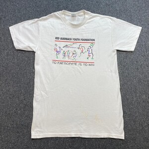 May include: A white short-sleeve t-shirt featuring a graphic print with the text RED AUERBACH YOUTH FOUNDATION and TO PARTICIPATE IS TO WIN. The design includes colorful line art illustrations of various athletes engaged in different sports activities.