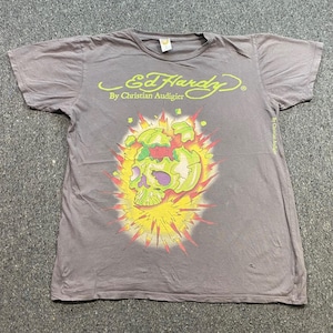 May include: A grey short-sleeve t-shirt featuring the Ed Hardy by Christian Audigier logo in yellow text at the top. The center graphic displays a stylized lime green skull surrounded by a bright yellow and red sunburst explosion design.