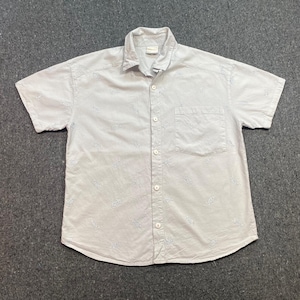 May include: A light-colored short-sleeve button-down shirt featuring a repeating pattern of small embroidered fish. The shirt includes a standard collar, a single chest pocket on the left side, and white buttons running down the center front placket.
