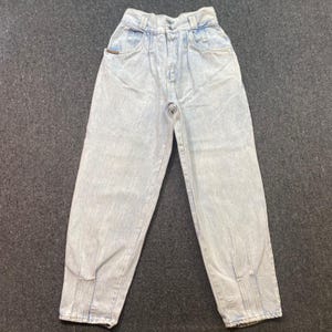 May include: High-waisted light wash denim jeans featuring a relaxed fit, tapered legs, and a pleated front design. These pants include a button and zipper fly closure, belt loops, and side pockets with a small brown leather-like brand tag detail.