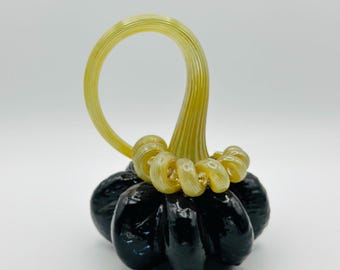 May include: A handcrafted glass pumpkin with a glossy black body and a yellow stem. The stem curves into a loop, and the base of the stem has decorative swirls. The pumpkin is a decorative item.
