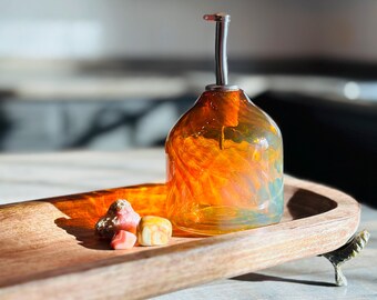 May include: An orange and yellow glass oil bottle with a stainless steel spout, presented on a wooden serving tray. The tray holds small, colourful candies and has a decorative metal foot. The bottle is a decorative kitchen item.