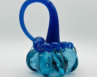 May include: A handcrafted glass sculpture in shades of blue. The piece features a rounded base with a textured surface and a looped handle. The sculpture's colour transitions from a light turquoise to a deep indigo.