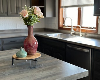 May include: A wooden tray with a black metal stand holds a pink and white marbled ceramic vase with pink roses and a teal ceramic vase. The tray is on a grey countertop in a kitchen.