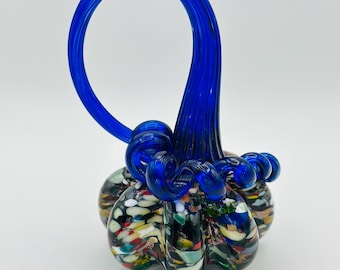 May include: A colourful glass pumpkin ornament with a striking blue handle. The pumpkin's body is a mix of speckled colours, and the handle is a solid, curved blue. A unique decorative item for the autumn season, perfect for home decor.