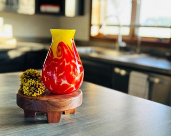 May include: A red and yellow glass vase with a swirl design sits on a wooden stand.  Yellow flowers are in front of the vase.