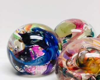 May include: Three glass paperweights with colourful swirls and bubbles inside. The paperweights are round and have a clear glass exterior.