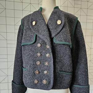 Puede incluir: Chaqueta de lana gris oscuro con diseño de doble botonadura. La chaqueta presenta botones dorados, ribetes verdes y cuello con solapas. Las mangas tienen puños doblados y dos bolsillos. La chaqueta se exhibe en un maniquí.