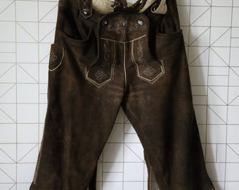 May include: Brown leather lederhosen with decorative embroidery and suspenders, hanging on a white hanger. The traditional Bavarian shorts feature detailed stitching and pockets, showcasing a classic design. The garment is displayed against a white geometric tile background.