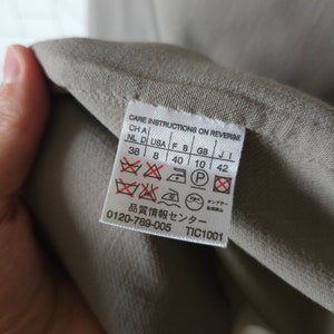 Op de afbeelding: Een beige waslabel met instructies op de achterkant. Het label bevat maatconversies voor verschillende landen, waaronder NL, D, USA, F, B, GB en J. Het bevat ook wasinstructies met symbolen voor wassen, strijken en chemisch reinigen. Het label bevat de tekst "品質情報センター" en "0120-789-005 TIC1001".