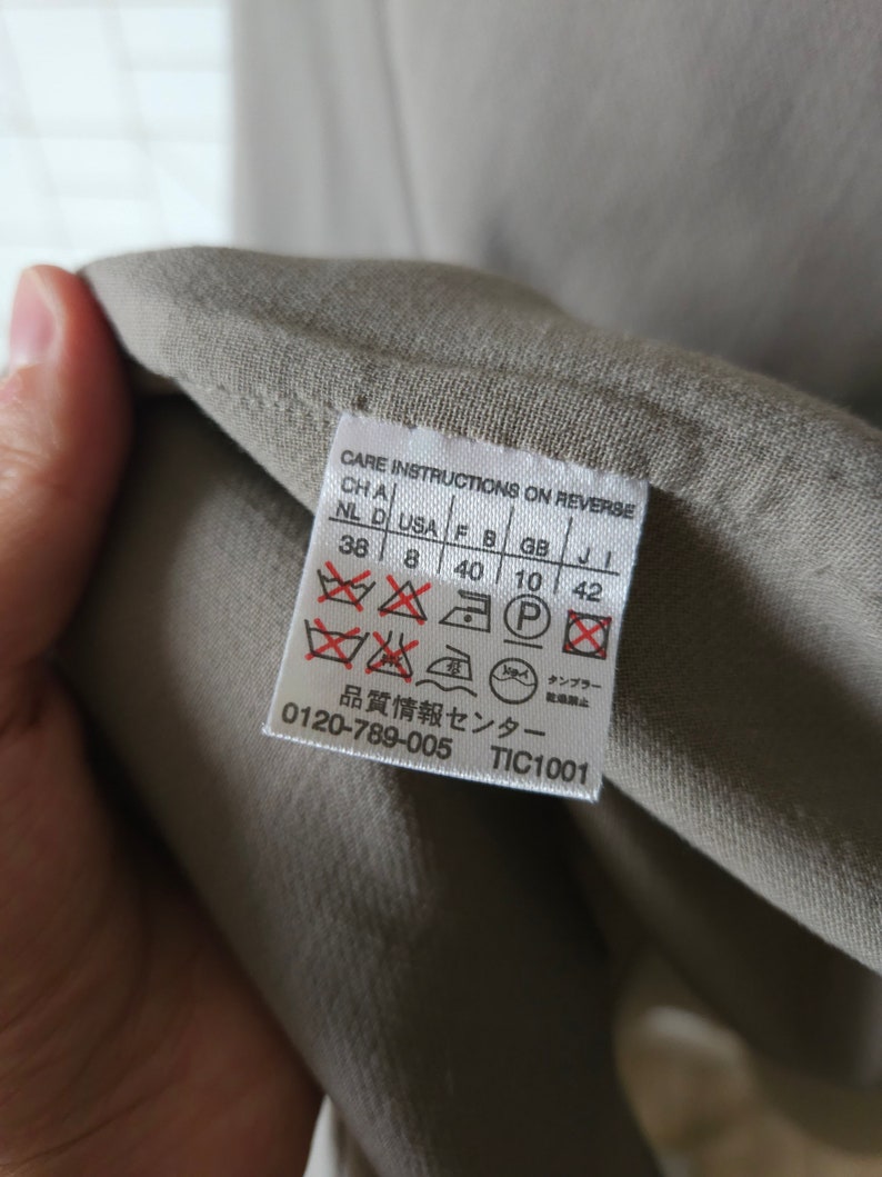 Op de afbeelding: Een beige waslabel met instructies op de achterkant. Het label bevat maatconversies voor verschillende landen, waaronder NL, D, USA, F, B, GB en J. Het bevat ook wasinstructies met symbolen voor wassen, strijken en chemisch reinigen. Het label bevat de tekst "品質情報センター" en "0120-789-005 TIC1001".