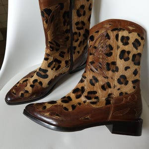 Könnte beinhalten: Ein Paar braune Cowboy-Stiefel aus Leder mit Leopardenmuster. Die Stiefel haben eine spitze Zehenpartie und einen niedrigen Absatz.