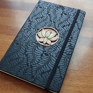 Puede incluir: Un diario negro con un patr&oacute;n floral y un adorno de madera en forma de flor de loto. El diario tiene un cierre el&aacute;stico negro.