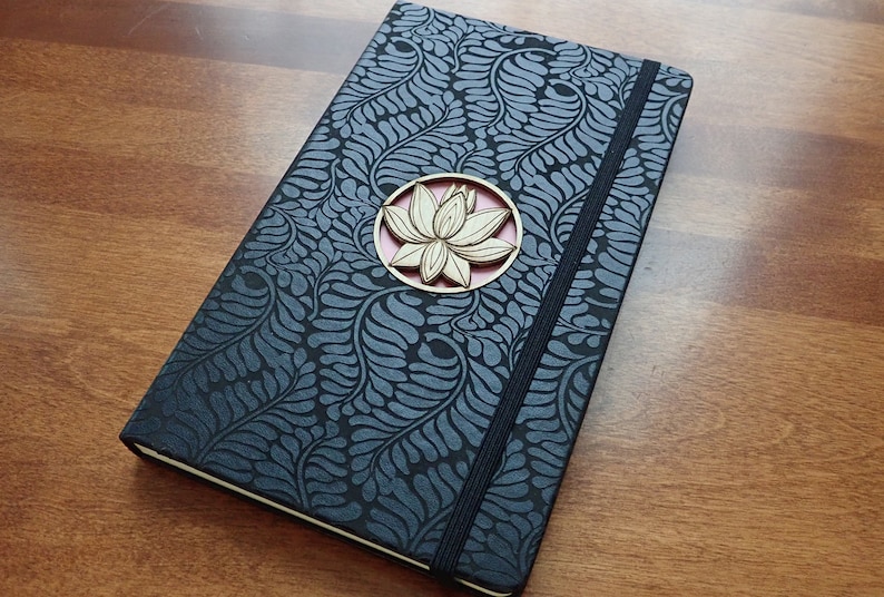 Puede incluir: Un diario negro con un patr&oacute;n floral y un adorno de madera en forma de flor de loto. El diario tiene un cierre el&aacute;stico negro.