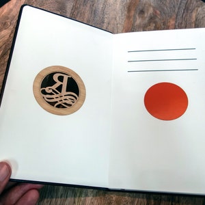 Puede incluir: Un cuaderno abierto con una cubierta negra. La p&aacute;gina izquierda tiene un c&iacute;rculo de madera con una letra R decorativa en su interior. La p&aacute;gina derecha tiene un c&iacute;rculo rojo.