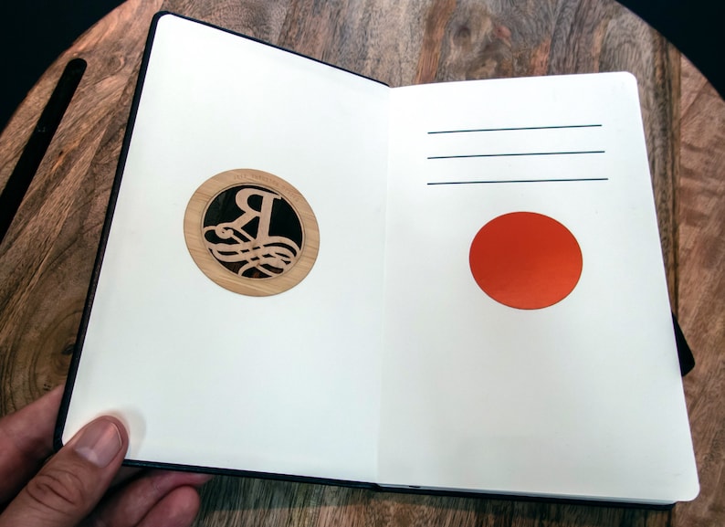 Puede incluir: Un cuaderno abierto con una cubierta negra. La p&aacute;gina izquierda tiene un c&iacute;rculo de madera con una letra R decorativa en su interior. La p&aacute;gina derecha tiene un c&iacute;rculo rojo.