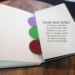 Puede incluir: Un cuaderno abierto con una cubierta negra y p&aacute;ginas blancas. El cuaderno tiene tres pegatinas de vinilo de colores en la parte posterior interior de la cubierta. Las pegatinas son de color morado, verde y rojo. El texto "Inside back folder: I'll always include a few extra color vinyl stickers in case you want to change colors. Just peel and stick on top of existing color on front page" est&aacute; impreso en la p&aacute;gina derecha.