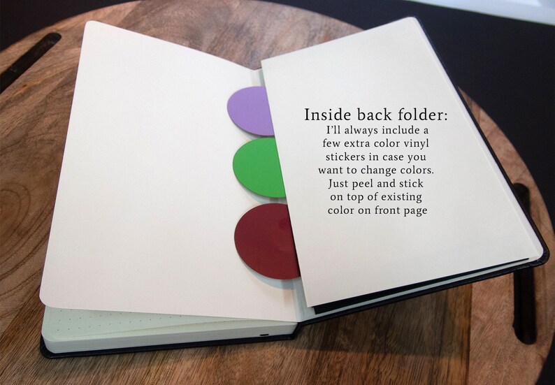 Puede incluir: Un cuaderno abierto con una cubierta negra y p&aacute;ginas blancas. El cuaderno tiene tres pegatinas de vinilo de colores en la parte posterior interior de la cubierta. Las pegatinas son de color morado, verde y rojo. El texto "Inside back folder: I'll always include a few extra color vinyl stickers in case you want to change colors. Just peel and stick on top of existing color on front page" est&aacute; impreso en la p&aacute;gina derecha.
