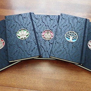 Puede incluir: Cinco diarios negros con diferentes acentos decorativos. Cada diario tiene un dise&ntilde;o circular con una flor de loto, un escarabajo, un &aacute;rbol, una mano de hamsa y otra flor de loto. Los diarios tienen un fondo estampado azul oscuro.