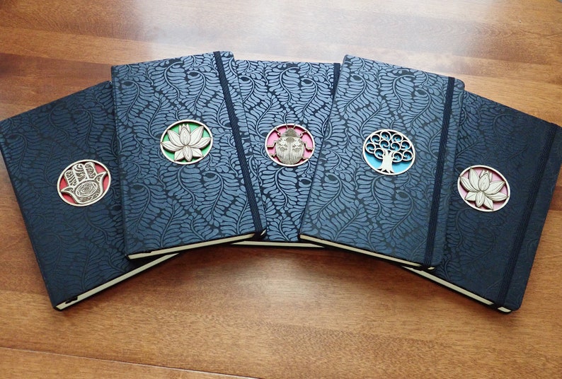 Puede incluir: Cinco diarios negros con diferentes acentos decorativos. Cada diario tiene un dise&ntilde;o circular con una flor de loto, un escarabajo, un &aacute;rbol, una mano de hamsa y otra flor de loto. Los diarios tienen un fondo estampado azul oscuro.