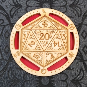 May include: A wooden D20 die with a red interior, featuring etched numbers and decorative symbols. The die is surrounded by a circular frame with additional etched details. The design is suitable for tabletop gaming.