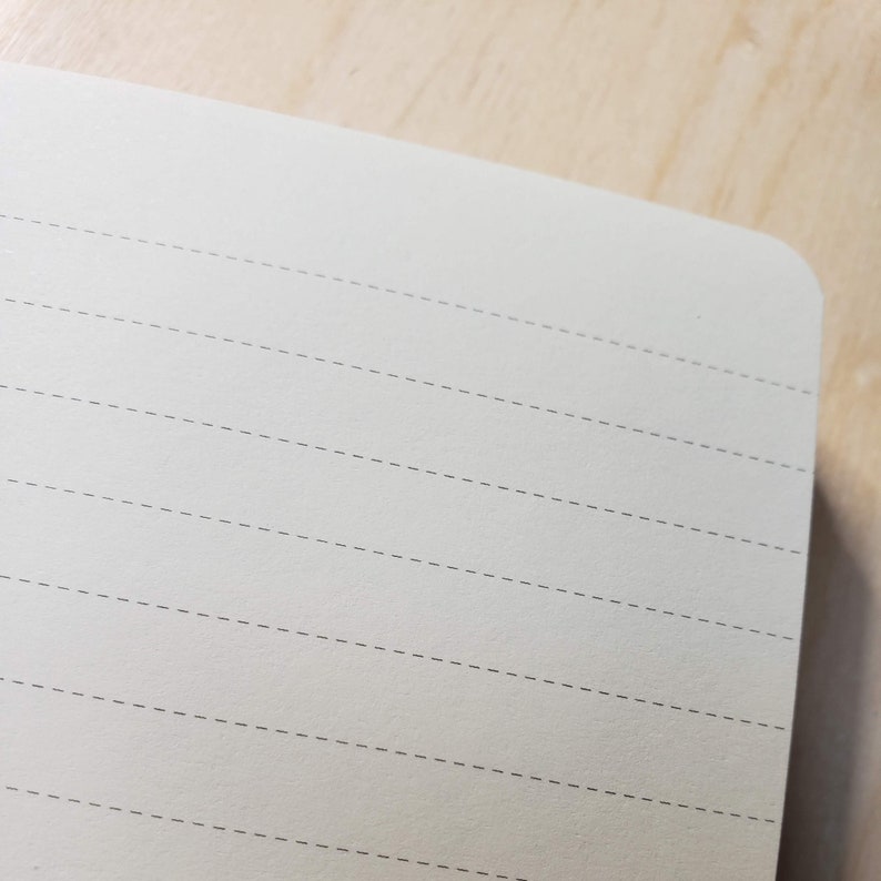 Puede incluir: Primer plano de un cuaderno blanco roto en blanco con p&aacute;ginas rayadas. Las l&iacute;neas son de un color gris sutil y est&aacute;n espaciadas uniformemente. El cuaderno est&aacute; hecho de un papel liso y texturizado.