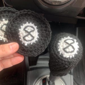 May include: Two black crocheted gear shift knobs with white embroidered number 8s. The knobs are in a car.