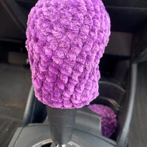 May include: A plush, purple crocheted shift knob cover. The cover is made of a soft, textured yarn and fits over the gear shift of a vehicle. The cover is a vibrant shade of purple and adds a touch of color to the car's interior.