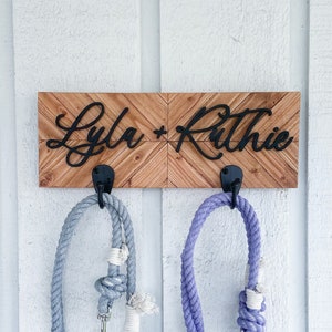 以下が含まれることがあります： 黒い筆記体で「Lyla」と「Ritchie」と書かれた木製の看板。看板のフックに、薄い青と紫のリードが2本掛かっている。