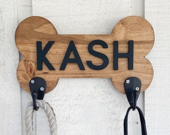 Op de afbeelding: Houten hondenbotvormige wandhaak met de naam "KASH" in zwarte letters. Twee riemen hangen aan de haken.